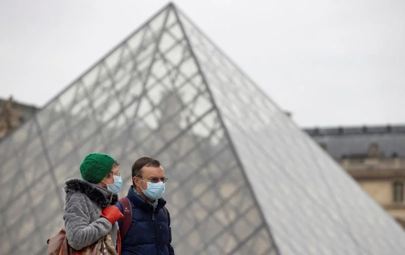 Two people walk next to the Louvre pyramid. 